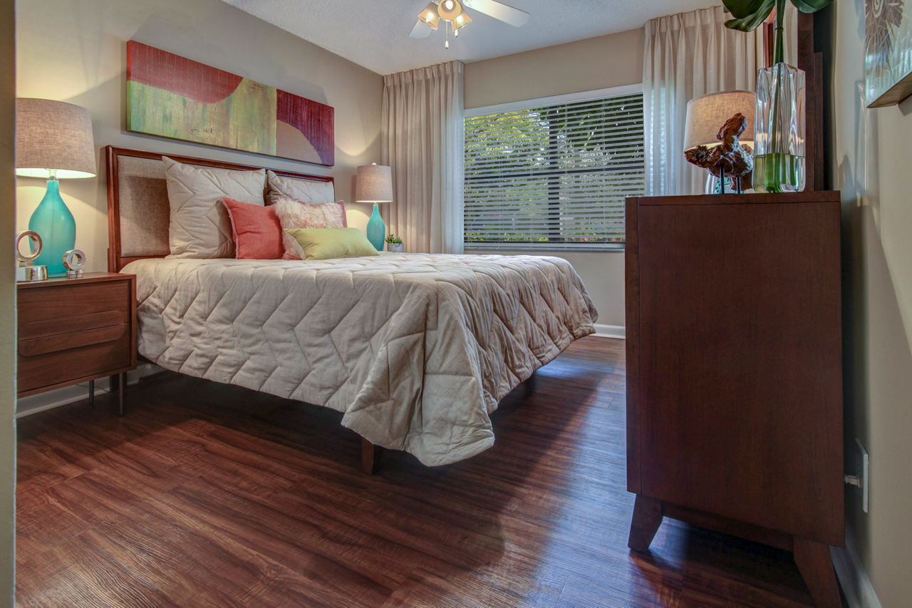 Cozy bedroom featuring wooden floors, elegant lamps, and sunlight from the window at The Plantation Colony Apartments