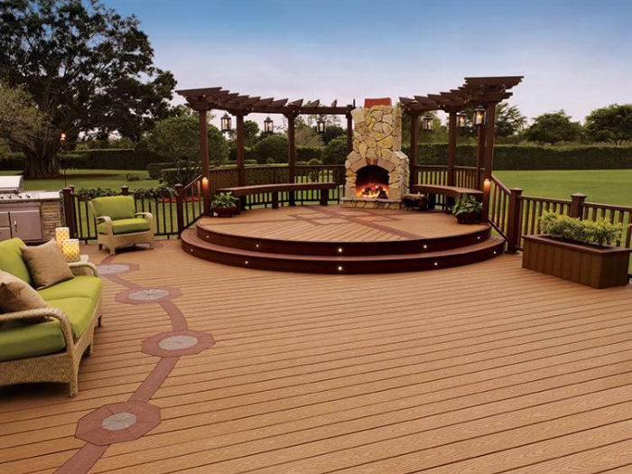 A large wooden deck features a circular raised platform with a stone fireplace and pergola. Comfortable green cushioned seating is arranged around the deck, creating a cozy outdoor living space. The setting is surrounded by lush grass and trees, with a clear sky overhead. The deck includes decorative inlays and built-in planters for added visual interest.