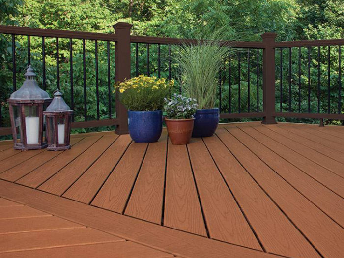 A spacious wooden deck is shown with several potted plants arranged along the railing. Two decorative lanterns are placed on the left side, adding a cozy touch to the outdoor setting. The deck overlooks lush green trees, creating a peaceful and inviting atmosphere. The scene is well-lit, highlighting the warm brown tones of the deck boards.