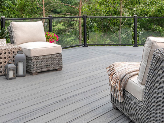 A spacious outdoor patio features two cushioned wicker lounge chairs and a woven side table, set against a backdrop of lush green trees. The deck has a sleek, gray wood-like surface and is enclosed by a glass railing. Decorative lanterns and a cozy throw blanket add a touch of comfort and style, while a potted plant brings color to the serene setting.