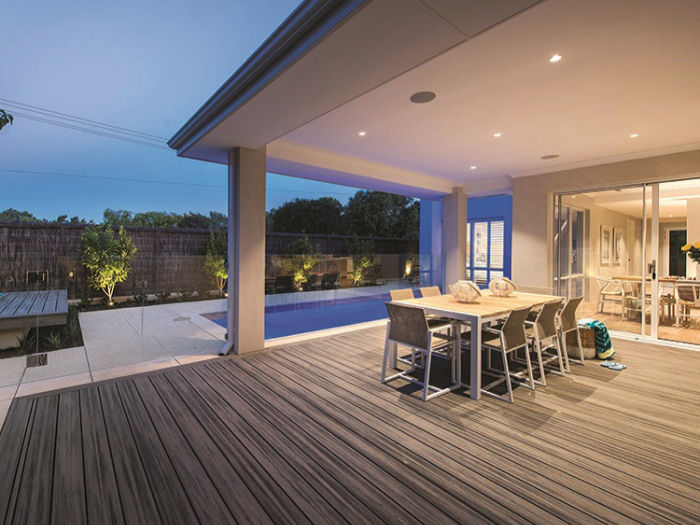 A contemporary outdoor dining space features a sleek table and chairs set on a wooden deck. The area opens up to a backyard with a swimming pool, softly lit in the evening. Large glass doors connect the patio to a stylish indoor dining room, creating a seamless indoor-outdoor living experience.