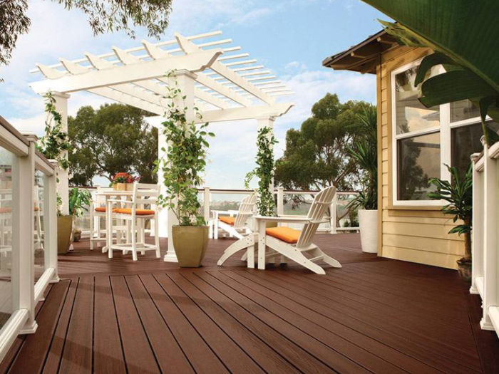 A spacious outdoor deck features rich brown wooden flooring and a white pergola adorned with climbing plants. White chairs with orange cushions and a dining table create a welcoming seating area. The setting is bright and airy, surrounded by lush greenery and adjacent to a yellow house. The overall mood is relaxed and inviting, perfect for outdoor gatherings.
