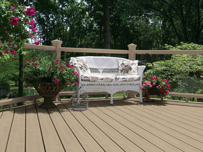 A cozy outdoor deck features a white wicker bench adorned with floral cushions, surrounded by lush potted plants and blooming flowers. The deck is made of light brown wood planks and is bordered by a simple railing. The setting is tranquil, with green trees in the background and bright pink and red flowers adding pops of color. The overall mood is peaceful and inviting, ideal for relaxation or enjoying nature.