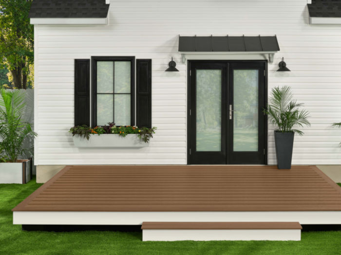 A contemporary white house features a brown wooden deck in the backyard, set against a backdrop of green grass. The house has black-framed double glass doors, a window with black shutters, and a flower box beneath the window. Potted plants and outdoor lighting fixtures add to the clean, inviting atmosphere.