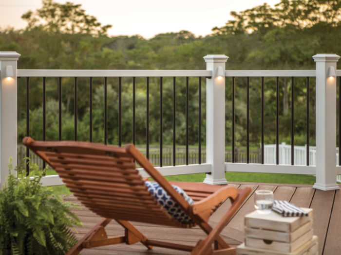 A wooden lounge chair with a patterned cushion sits on a wooden deck, facing a white and black railing. The setting is outdoors with lush greenery and trees in the background, suggesting a peaceful, natural environment. A small stack of books and a glass of water rest on a side table, enhancing the relaxed mood. Soft lighting on the railing posts adds a warm, inviting ambiance.