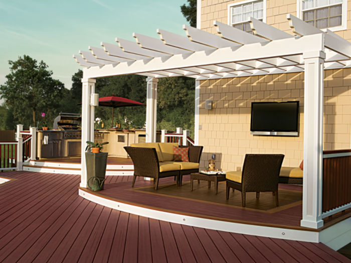 A spacious backyard deck features a white pergola over a cozy seating area with wicker chairs and a small table. The setting includes a mounted outdoor television and a built-in grill station in the background, ideal for entertaining. The deck has a rich reddish-brown wood finish, and the scene is set during daylight with clear skies. No visible people or text are present in the image.
