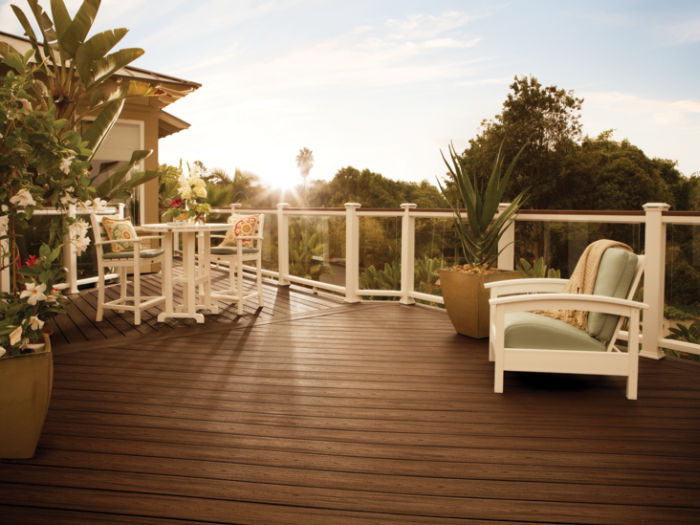 A spacious wooden deck is shown at sunset, featuring white outdoor furniture including a cushioned chair and a small dining set. Lush green plants in pots line the perimeter, and a glass railing offers a view of surrounding trees. The scene is bathed in warm, golden light, creating a tranquil and inviting atmosphere.