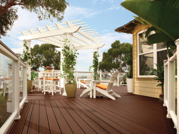 A spacious wooden deck features a white pergola adorned with climbing plants and a set of white outdoor furniture. The setting is bright and inviting, with lush greenery and trees surrounding the area. The deck includes glass railings and comfortable chairs with orange cushions, creating a relaxed and welcoming atmosphere.